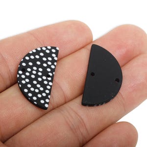 May include: Two black, semi-circular pendants. One is patterned with white polka dots, the other is plain black. Each pendant has two small holes for threading.