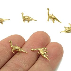 May include: Gold-coloured dinosaur charms in various poses. These small, detailed pendants feature different dinosaur species, ideal for jewellery making or crafting projects. The charms are shown against a white background.