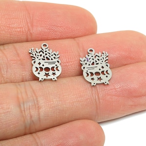 May include: Two small, silver-colored cauldron charms with a witchy theme. Each charm features a cauldron shape with crescent moons, stars, and foliage details. The charms have a small loop at the top for attaching to jewelry or other crafts.