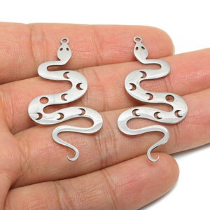 May include: Two silver snake charms. Each charm has a loop at the top and a curved tail. The snake's body features a series of crescent moon cutouts. These charms are likely for jewellery making or crafting.