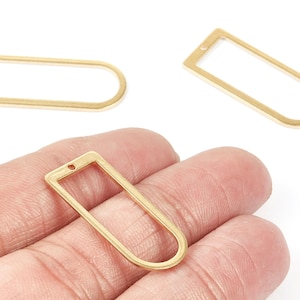 Puede incluir: Tres colgantes dorados con marco abierto. Uno se sostiene en una mano, mientras que otros dos están dispersos sobre una superficie blanca. Cada colgante tiene una parte superior rectangular con una parte inferior redondeada y un pequeño agujero para ensartar.