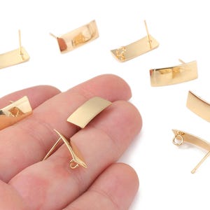 May include: Gold-coloured rectangular stud earrings with a curved design. The earrings have a post and a small loop for attaching to the ear. Several earrings are scattered on a white background.