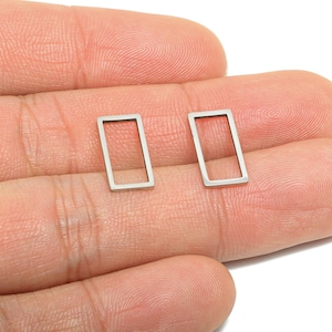 Puede incluir: Dos pequeños pendientes rectangulares plateados. Cada pendiente presenta un diseño de marco abierto y sencillo. Los pendientes se muestran sobre un fondo color piel, destacando su estilo minimalista y su potencial para el uso diario.