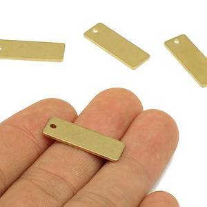 May include: Several small, rectangular brass charms with a hole at one end are displayed. One charm is held in a hand, while others are scattered in the background. The charms have a smooth, matte finish and are likely used for jewelry making or crafting.