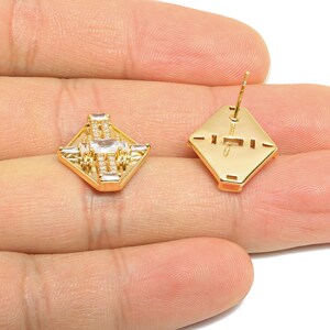 May include: Gold-coloured stud earrings with a geometric design. Each earring has a square shape with a central arrangement of clear, rectangular stones and smaller accent stones. The earrings are displayed against a neutral background.