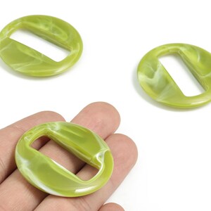 Puede incluir: Tres hebillas redondas de resina verde oliva y blanco veteado. Una hebilla se sostiene en la mano. Las hebillas tienen una abertura rectangular, ideales para proyectos de manualidades. Diámetro aproximado de 3 cm.