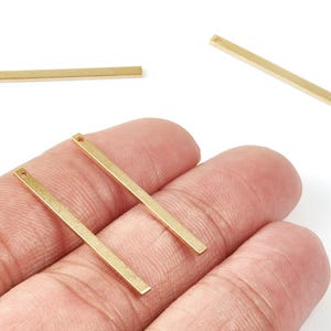 Puede incluir: Se muestran varios dijes rectangulares dorados con un pequeño agujero en un extremo. Los dijes se muestran sobre un fondo blanco, algunos sostenidos en una mano. Estos componentes para la fabricación de joyas son ideales para crear collares y pendientes.
