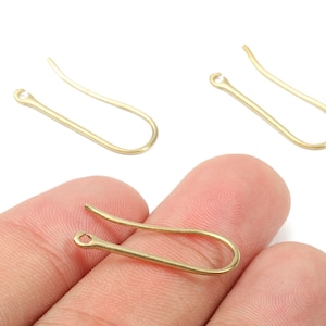 May include: Three gold-colored earring hooks are displayed against a white background. One hook is held between fingers. The hooks have a curved design with a small loop at one end and a curved hook at the other. Jewelry making supplies.