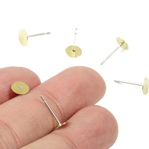May include: A collection of gold-colored earring posts with flat, round pads. The posts are silver-colored, and the pads are a bright, shiny gold. Several posts are scattered on a white background, with one held between fingers.