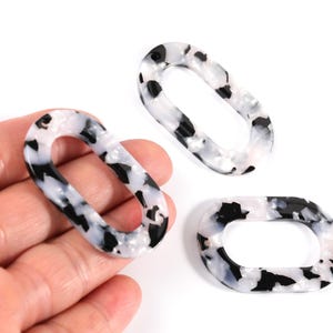 Peut inclure: Trois maillons ovales en résine avec un motif moucheté noir et blanc. Les maillons sont dans différentes orientations, dont un tenu dans une main. Les maillons sont de couleur neutre et peuvent être utilisés pour la fabrication de bijoux ou d'autres travaux manuels.