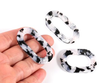 May include: Three oval-shaped resin links with a black and white speckled pattern. The links are in various orientations, with one held in a hand. The links are a neutral color and can be used for jewelry making or other crafts.