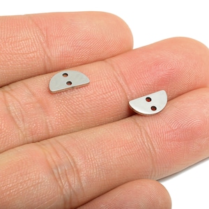 Puede incluir: Dos pequeños botones plateados en forma de semicírculo con dos agujeros cada uno. Los botones se muestran en la mano de una persona.