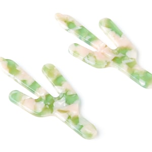 May include: Two green and pink cactus-shaped hair clips. The clips are made of a marbled plastic material.