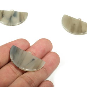 Puede incluir: Tres colgantes semicirculares de resina veteada en tonos grises y crema. Cada colgante tiene un pequeño agujero en la parte superior para ensartar. Los colgantes se muestran sobre un fondo blanco.