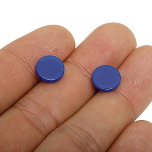 May include: Two small, round, cobalt blue objects are displayed. The objects are smooth and shiny, with a slightly raised edge. The items are held in a person's hand, against a white background. The objects appear to be buttons or decorative elements.