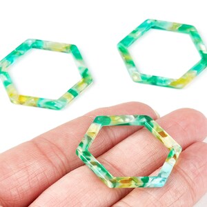 May include: Three open-center, hexagon-shaped beads. The beads have a marbled pattern of green, yellow, and white. One bead is held in a hand. The beads are made of a translucent material.