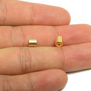 May include: Two small, gold-colored cylindrical beads. One bead is positioned on its side, while the other stands upright, revealing a small hole. The beads are displayed against a neutral background.