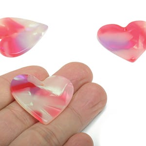 May include: Three heart-shaped pendants with a marbled effect. The pendants feature shades of pink, red, and white. One pendant is held in a hand. The pendants are likely for jewellery making or crafting.