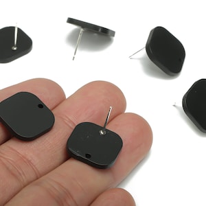 May include: Black square stud earrings with rounded corners. Each earring has a silver post and a small hole. Several earrings are scattered on a white background.