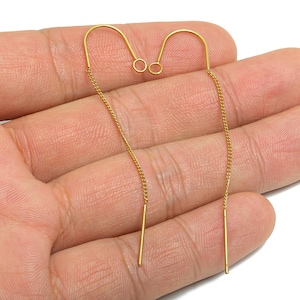 May include: A pair of gold-colored threader earrings. Each earring features a curved top with a small loop, a delicate chain, and a straight bar at the end. The earrings are displayed against a light background.