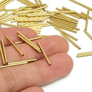 Può includere: Primo piano di numerosi piccoli tubi cilindrici in ottone. Alcuni tubi sono tenuti in mano, mentre altri sono sparsi su una superficie bianca. I tubi sono lunghi circa 1,27 cm e hanno un centro cavo.