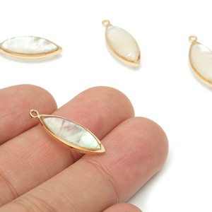 May include: Four marquise-shaped jewelry pendants. Each pendant features a pearlescent, iridescent center stone set in a gold-colored bezel. One pendant is held by a hand. The pendants have a small loop at the top for attaching to a chain or other jewelry component.