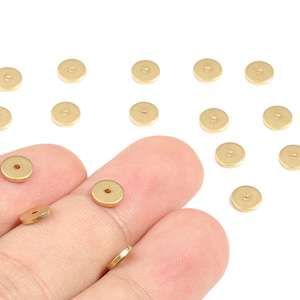 Puede incluir: Una colección de pequeños discos redondos de metal dorado con un agujero central. Algunos de los discos se sostienen entre los dedos, mientras que otros están dispersos sobre una superficie blanca. Probablemente hallazgos o componentes de joyería.