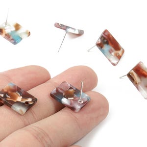 May include: Rectangular stud earrings with a marbled pattern in shades of brown, white, and blue. The earrings have a post backing. Several earrings are scattered on a white background.