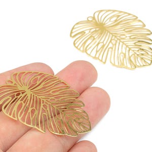 May include: Two gold-colored, openwork metal leaf charms. One is held in a hand, and the other is positioned above it. The charms are shaped like Monstera leaves, with intricate vein details. These charms are suitable for jewelry making.
