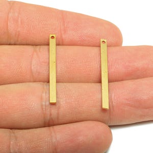 May include: Two small, rectangular, gold-colored metal bar pendants with a hole at the top. The pendants are held in a person's hand. These jewelry findings are ideal for creating earrings, necklaces, or other handmade jewelry.