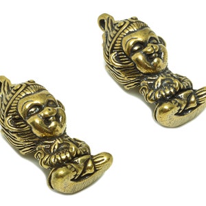 Puede incluir: Dos amuletos de metal de color dorado que representan una pequeña figura sentada con una corona y una expresión traviesa. Los amuletos son detallados y tienen un estilo asiático tradicional.