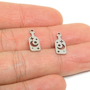 May include: Two small, silver-colored bottle charms. Each charm features a bottle shape with a crescent moon and star cut-out design. The charms are held in a person's hand against a white background.