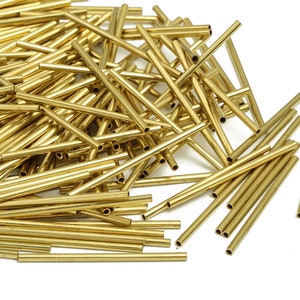 Puede incluir: Un montón de tubos de latón de color dorado, esparcidos sobre una superficie blanca. Los tubos son de varias longitudes y todos son huecos.