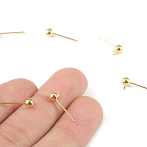 May include: Gold-coloured earring posts, each with a small ball and a loop for attaching charms. The posts are scattered on a white background. The earring posts are suitable for creating custom jewellery.