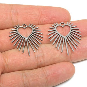 Könnte beinhalten: Zwei herzförmige Anhänger in Silber mit Sonnenstrahlendesign. Jeder Anhänger hat einen Herz-Ausschnitt in der Mitte und nach außen gerichtete Linien. Die Anhänger werden vor einer Hand gezeigt.