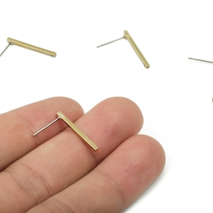 May include: Gold-colored bar earrings with silver posts. The earrings are rectangular and have a minimalist design. Several earrings are scattered on a white background, with one held in a hand.