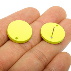May include: Two round, bright yellow earring studs. One stud has a post and backing, the other has a small hole. The earrings are held in a hand.