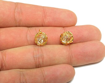 May include: Two gold-colored, openwork sphere pendants with a clear, faceted stone inside. The pendants are held in a hand against a white background. The pendants have a small loop at the top for attaching to a chain or earring hook.