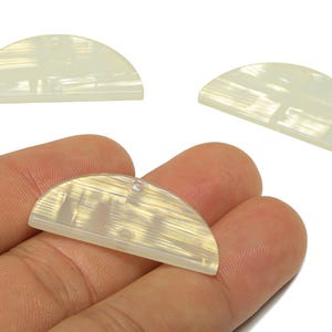May include: Three translucent, half-circle shaped beads with a pearlescent finish. One bead is held in a hand, while two others are positioned above. Each bead has a small hole for stringing.