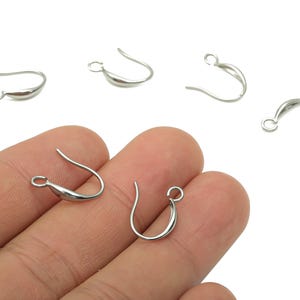 Könnte beinhalten: Silberfarbene Ohrhaken mit geschwungenem Design. Mehrere Haken sind auf weißem Hintergrund verstreut, zwei werden von einer Hand gehalten. Die Haken haben oben eine kleine Schlaufe und einen geschwungenen, tropfenförmigen Körper.