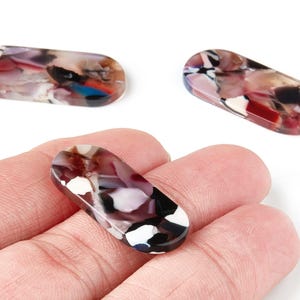 May include: Three oval-shaped resin beads with a marbled pattern of red, white, black, and blue. One bead is held in a hand. The beads have a glossy finish and are likely for jewelry making.