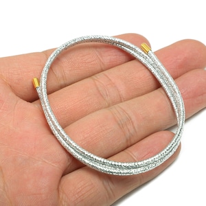 Puede incluir: Una pulsera plateada con superficie texturizada y extremos dorados. La pulsera circular está hecha de múltiples hebras. La joya se muestra en una mano sobre un fondo blanco.