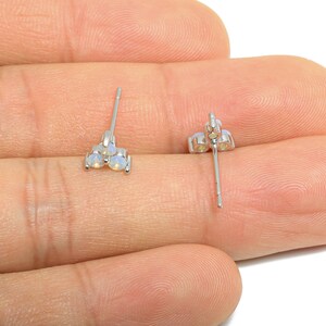 Puede incluir: Un par de pendientes de plata con tachuelas, cada uno con tres pequeñas piedras redondas y opalinas. Los pendientes se muestran sobre un fondo neutro, destacando su delicado diseño y sutil brillo. Los postes son rectos y delgados.