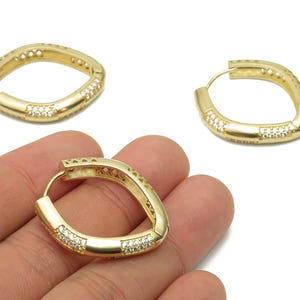 Op de afbeelding: Goudkleurige oorringen met een uniek, licht gebogen ontwerp. Elke oorring heeft een rij kleine, heldere edelstenen en decoratieve uitsparingen. De oorbellen worden getoond tegen een witte achtergrond.