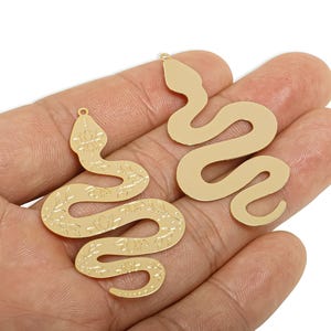 Può includere: Due ciondoli a forma di serpente color oro. Un ciondolo presenta un motivo floreale inciso lungo il corpo, mentre l'altro ha un design liscio e uniforme. Entrambi hanno un piccolo anello nella parte superiore per essere appesi. Ideale per la creazione di gioielli.