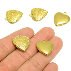 Könnte beinhalten: Goldfarbene herzförmige Medaillons. Einige Medaillons haben florale Designs, andere sind glatt. Jedes Medaillon hat oben eine kleine Schlaufe zum Befestigen an einer Kette oder anderem Schmuck. Die Medaillons sind auf weißem Hintergrund.