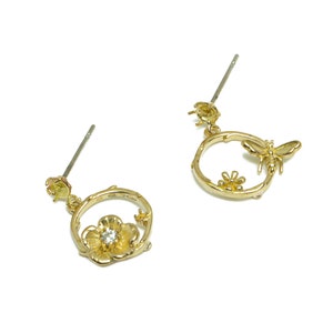May include: A pair of gold-tone earrings with a floral design. Each earring features a circular frame with a flower and a bee. The flower has a clear rhinestone in the center.