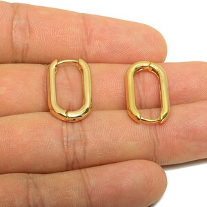 May include: Two gold-coloured, oval-shaped hoop earrings. The earrings are a shiny gold colour and have a simple, modern design. The earrings are shown against a neutral background.