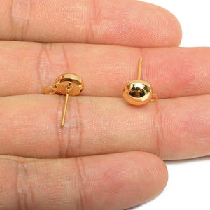 May include: Two gold-colored stud earrings with a round, polished design. Each earring has a post for insertion and a small loop at the base of the round element. The earrings are shown against a skin-toned background.