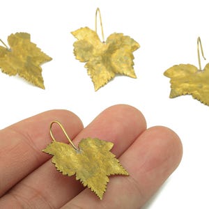 May include: Gold-colored leaf-shaped earrings. The earrings have a textured surface and a hook closure. The image shows three earrings, with one held in a hand. The earrings are made of metal and have a natural leaf design.